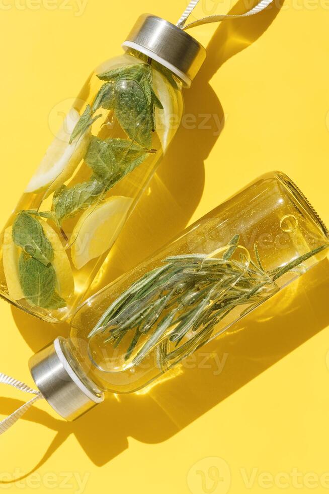 Top view closeup two glass bottles, water drink detox with lemon and mint leaves, and rosemary, at sunlight on yellow color background. Infused water, wellness trend. Refreshing summer drink photo