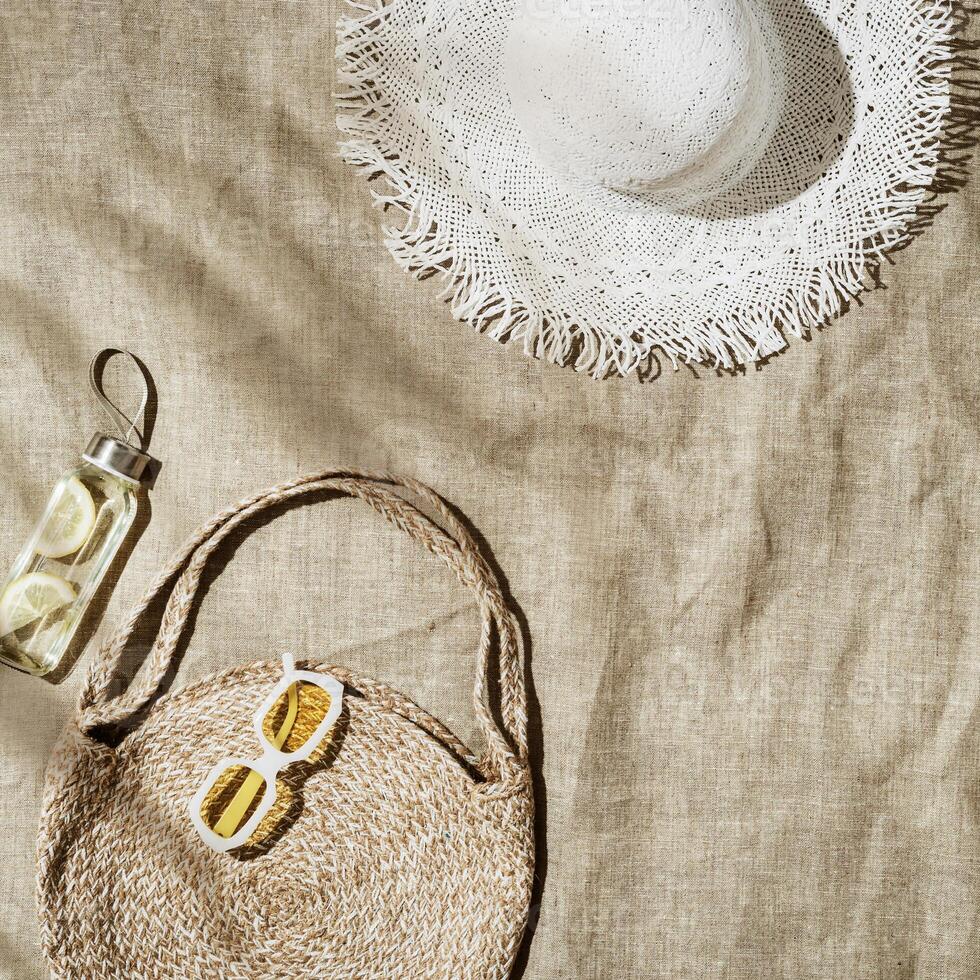 Beach still life at summer sun, fashion jute beach bag, detox lemon water, sun hat, sunglasses on beach towel, summer relax concept, image of modern stylish relaxing on beach, top view, space photo