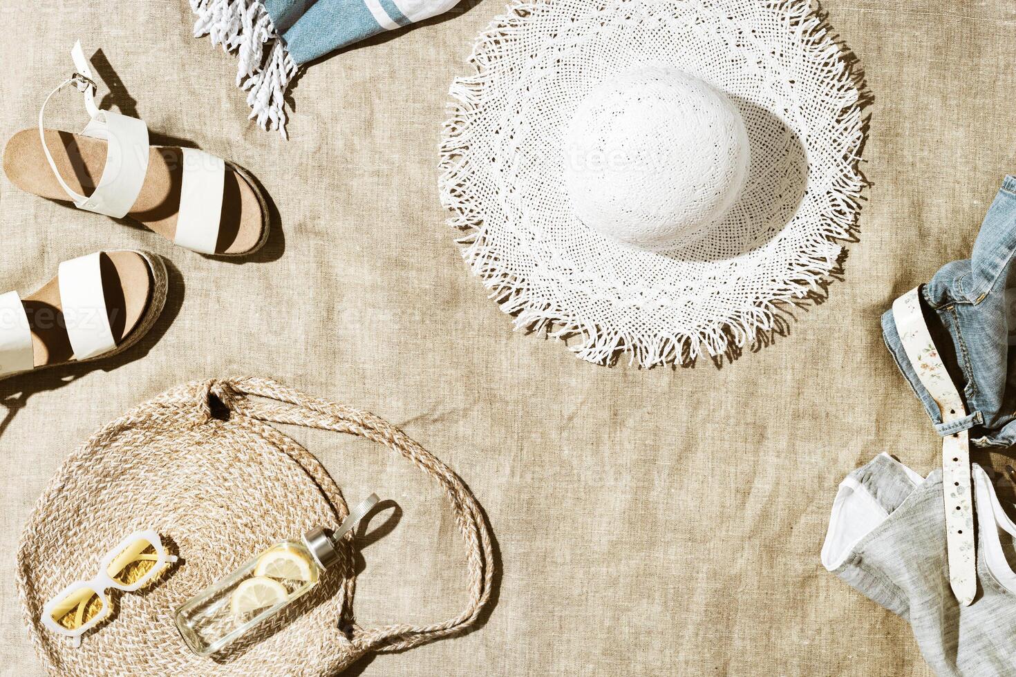 Summer flat lay fashion casual clothes, beach bag, trendy yellow sunglasses, white sandals, sun hat, shorts, lemon water in bottle on beach towel at sunlight, relaxing on beach, summer mood photo