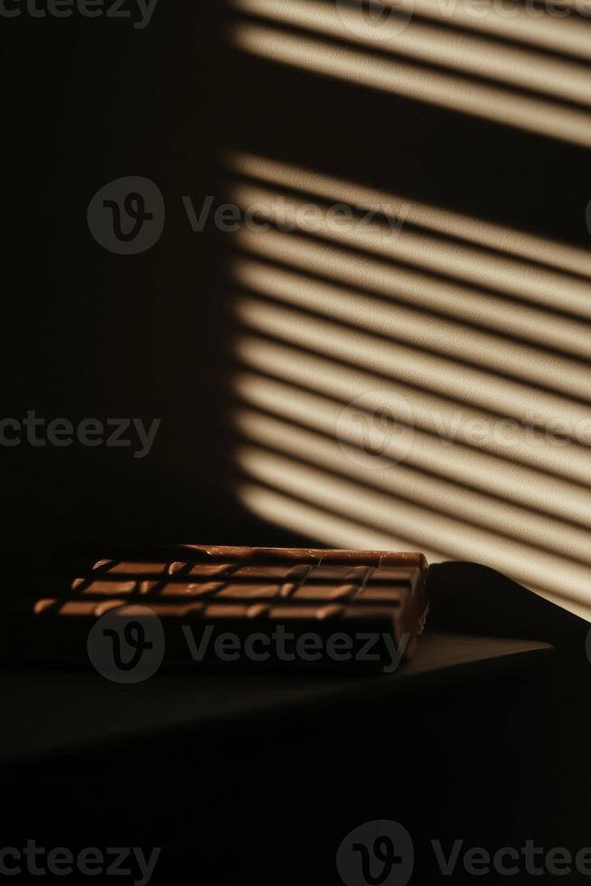 A captivating image of a brown chocolate bar resting on a table, illuminated by soft sunlight filtering through blinds, creating beautiful shadows and patterns. photo