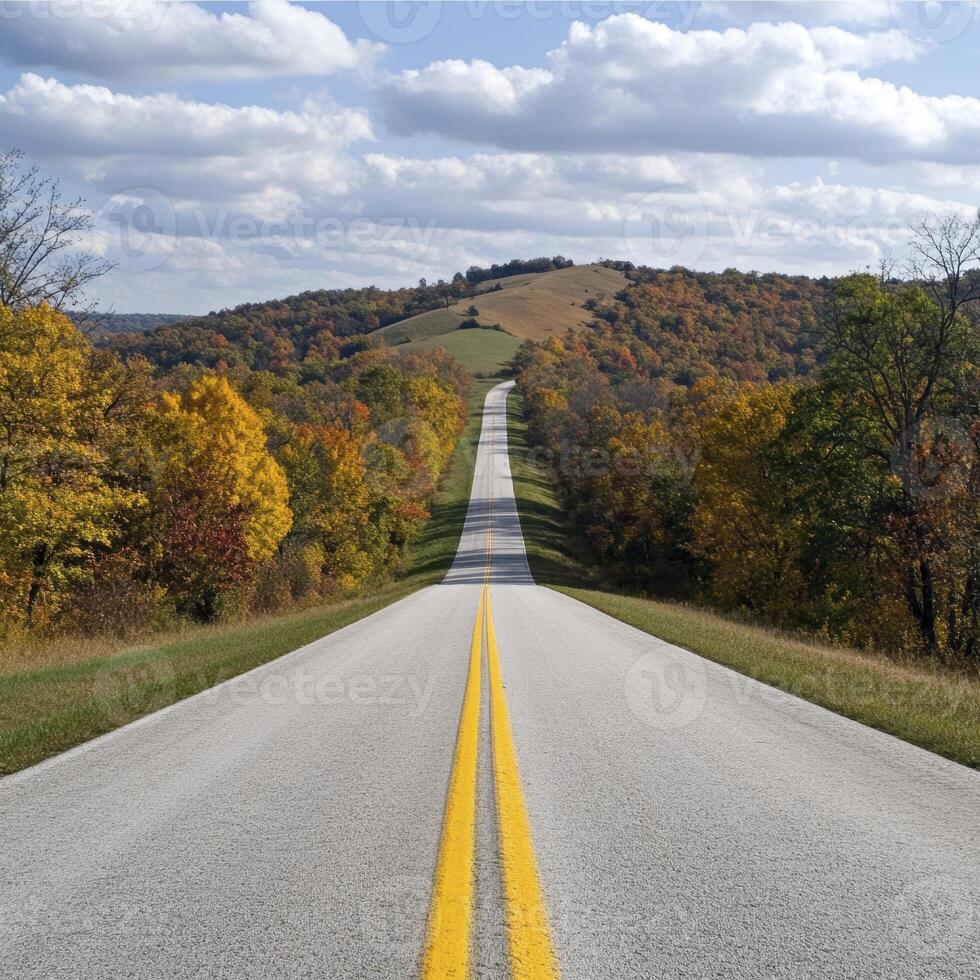 A picturesque country road stretches into the distance, flanked by vibrant autumn trees ablaze with color The yellow lines guide the eye towards a gentle, rolling hill in the horizon, topped with a photo