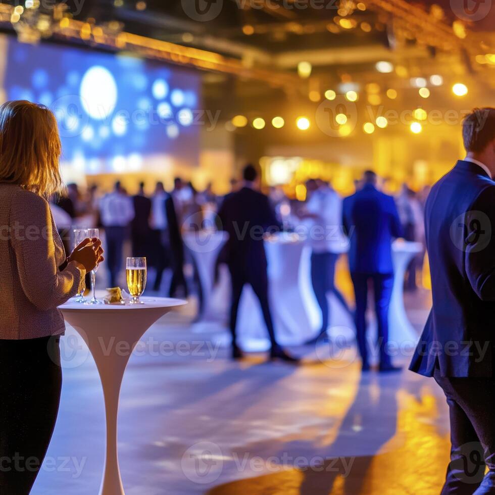 Amidst a soft, ethereal glow, an indoor corporate event unfolds, a symphony of blurred figures hinting at lively conversations and shared experiences The scene is bathed in warm light, casting an photo