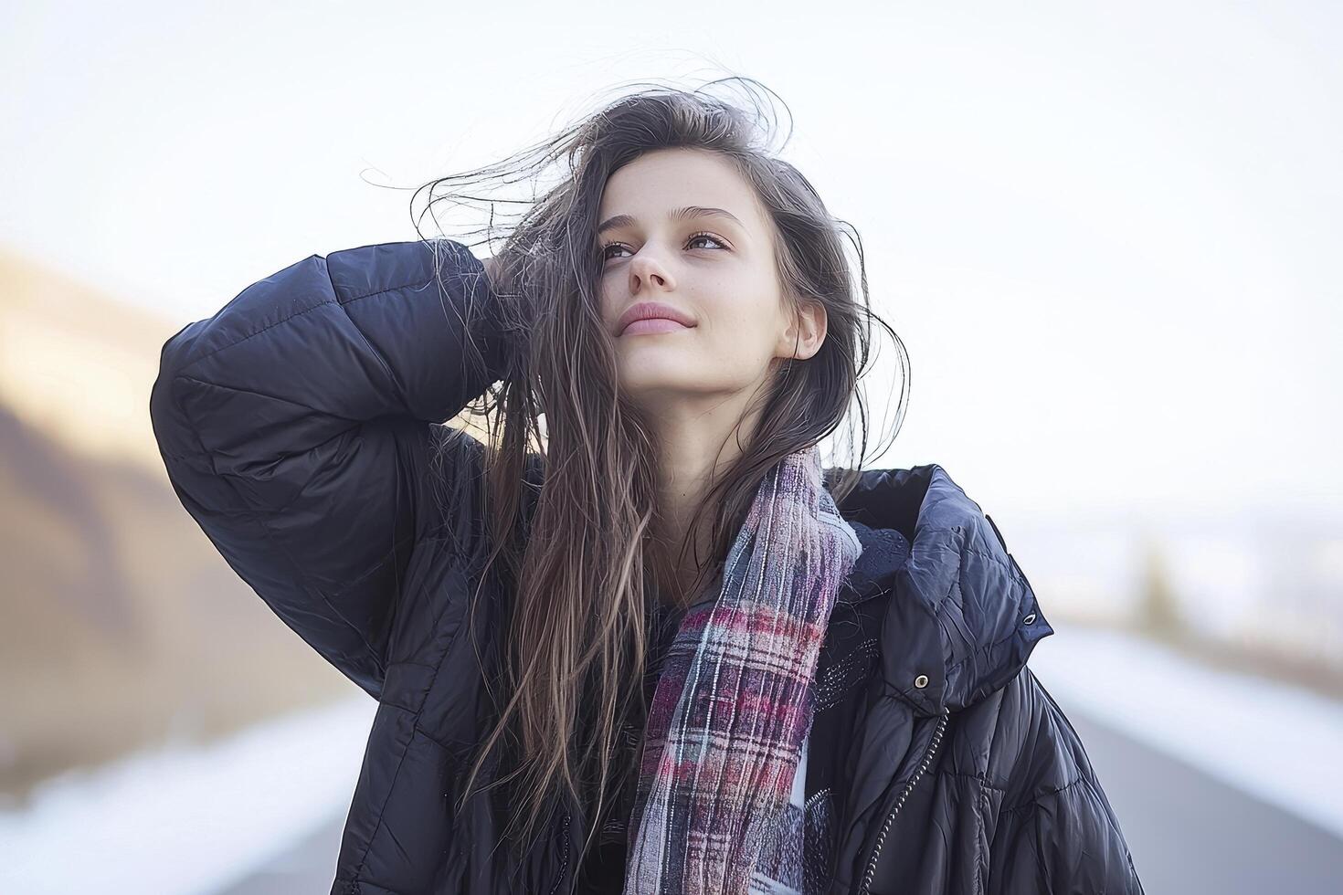 A serene portrait captures a young woman against a bright, slightly blurred backdrop Her long, wavy brown hair flows loosely, framing her face as she gazes upwards with a gentle smile She is dressed photo