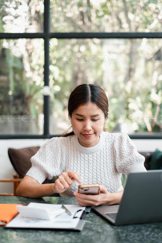 Young woman using a smartphone at a home office desk with a laptop and documents, enjoying a relaxed atmosphere with natural light. photo