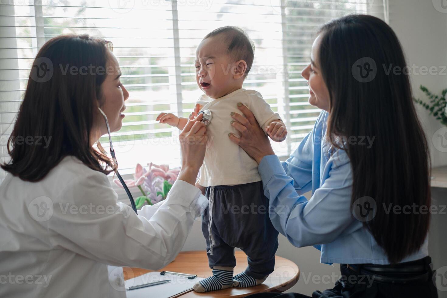 A pediatrician uses a stethoscope to examine a crying baby held by the mother in a well-lit medical office. photo