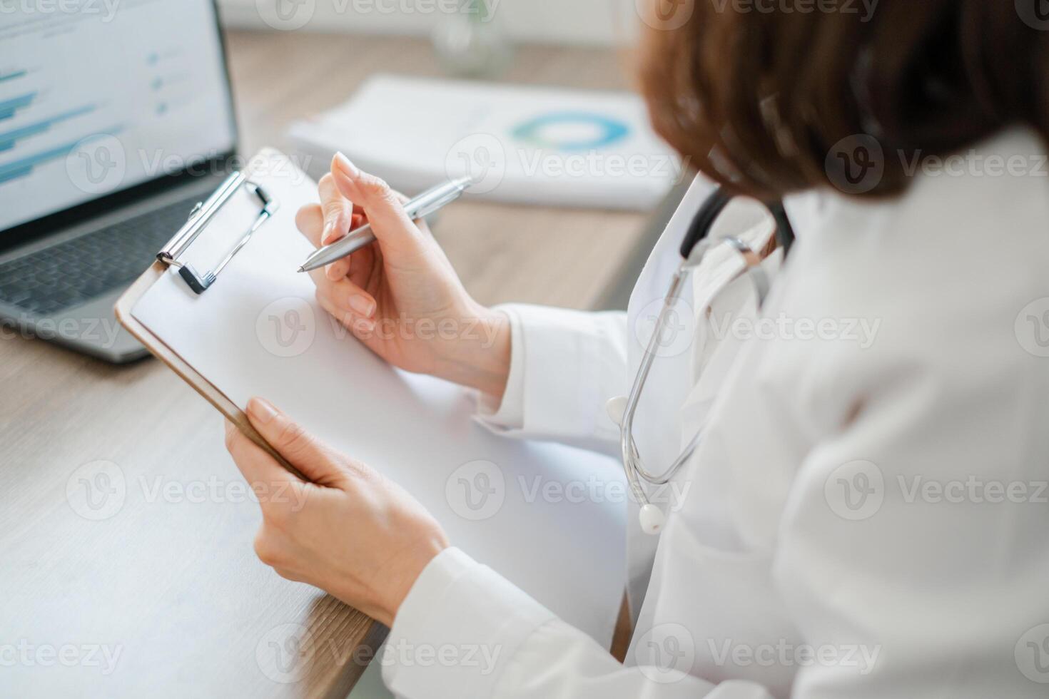 Doctor in white coat writing on clipboard with laptop and charts, symbolizing healthcare, professionalism, and medical documentation. photo