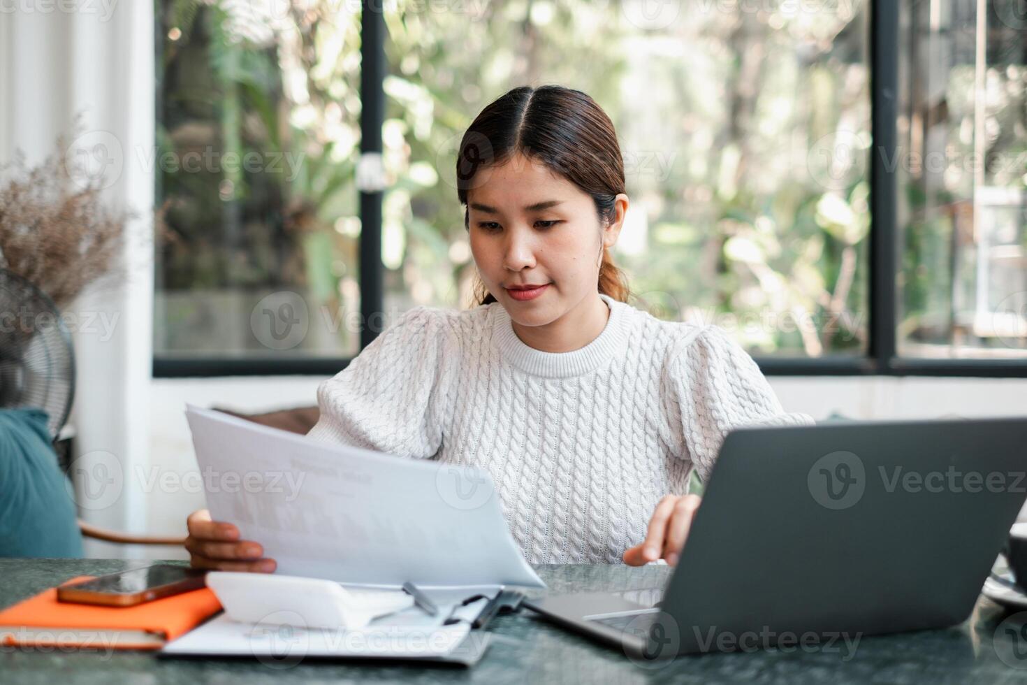 A focused young woman working from home, reviewing documents with a laptop in a bright, modern workspace. photo