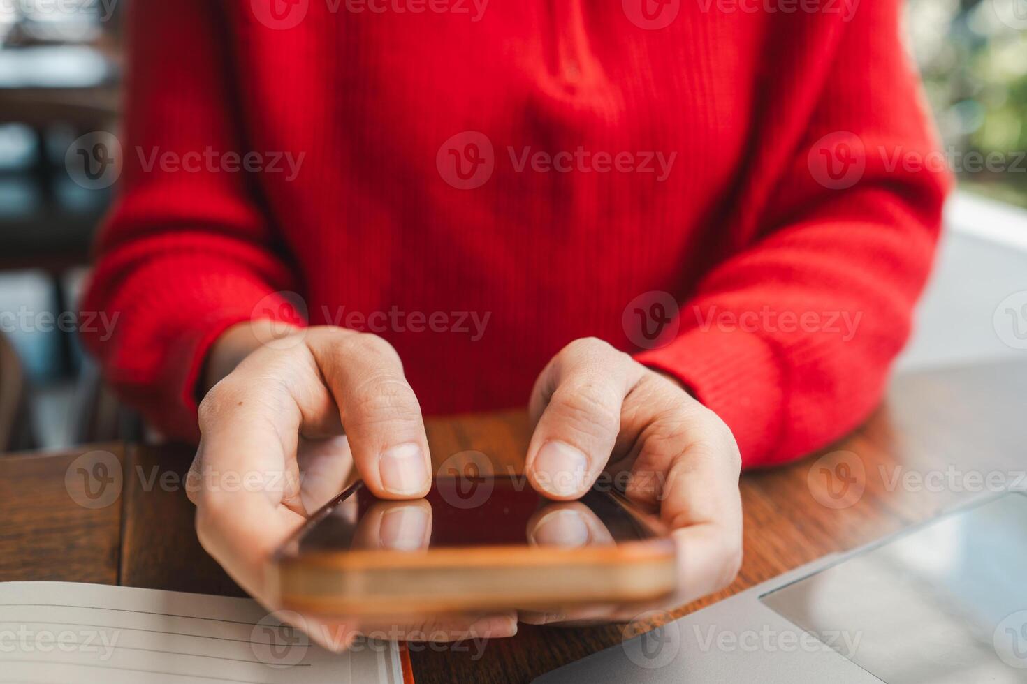 Hands holding a smartphone, wearing a red sweater, at a wooden table, capturing a moment of modern technology use. photo