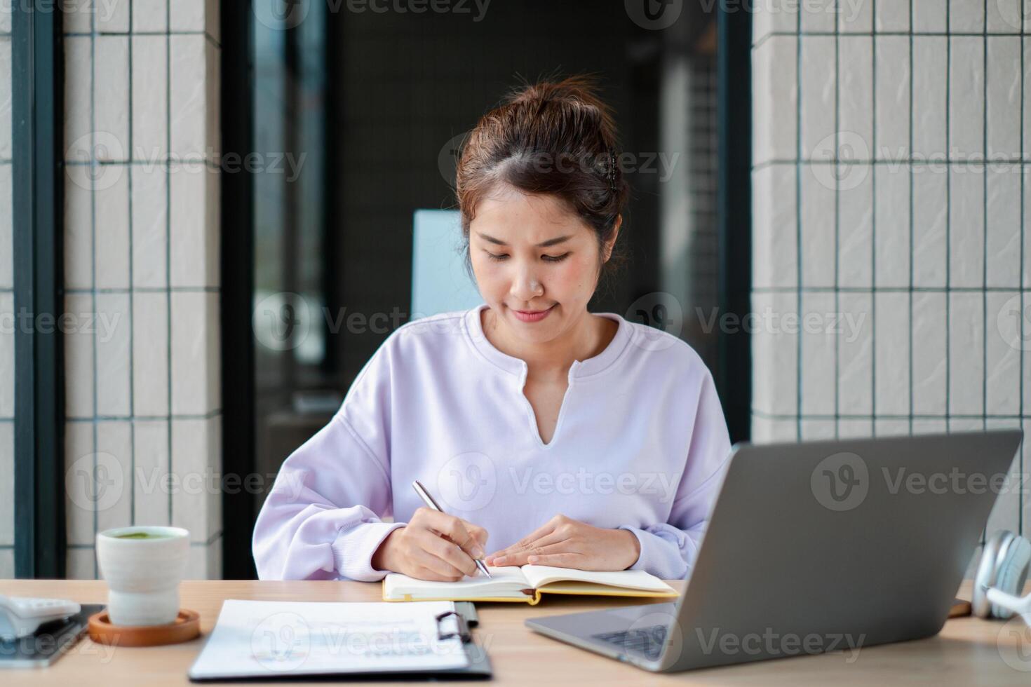 Woman concentrating on writing notes at a desk with a laptop, showcasing a productive work setting. photo