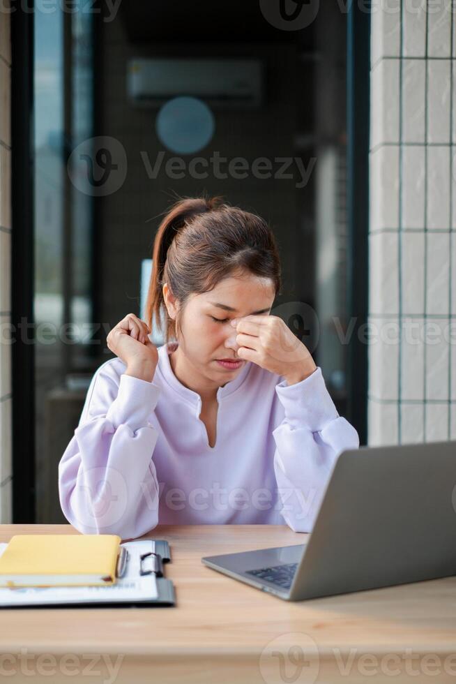 A woman in a white blouse appears stressed while sitting at a desk with a laptop and documents in a modern office environment. photo