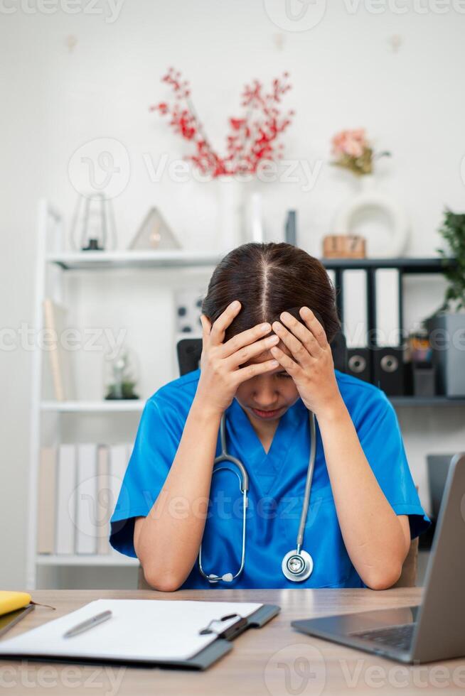A healthcare worker in blue scrubs appears stressed while sitting at a desk with medical documents and equipment in an office. photo