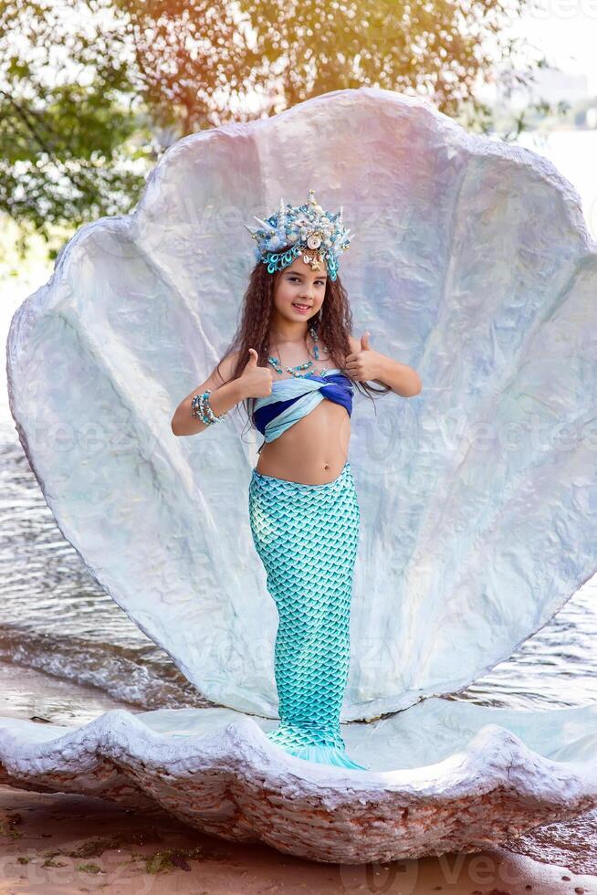 A beautiful girl in a mermaid costume stands outdoor in a large sea shell, photo