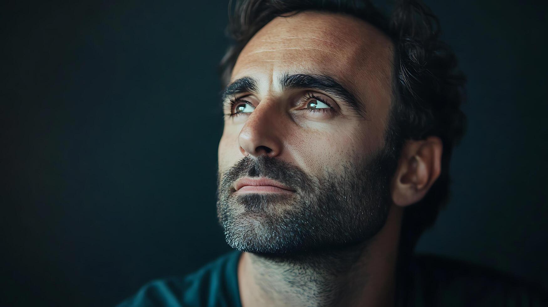 Pensive man gazes upward with thoughtful eyes He has a dark beard and hair against a dark background and hopeful expression photo