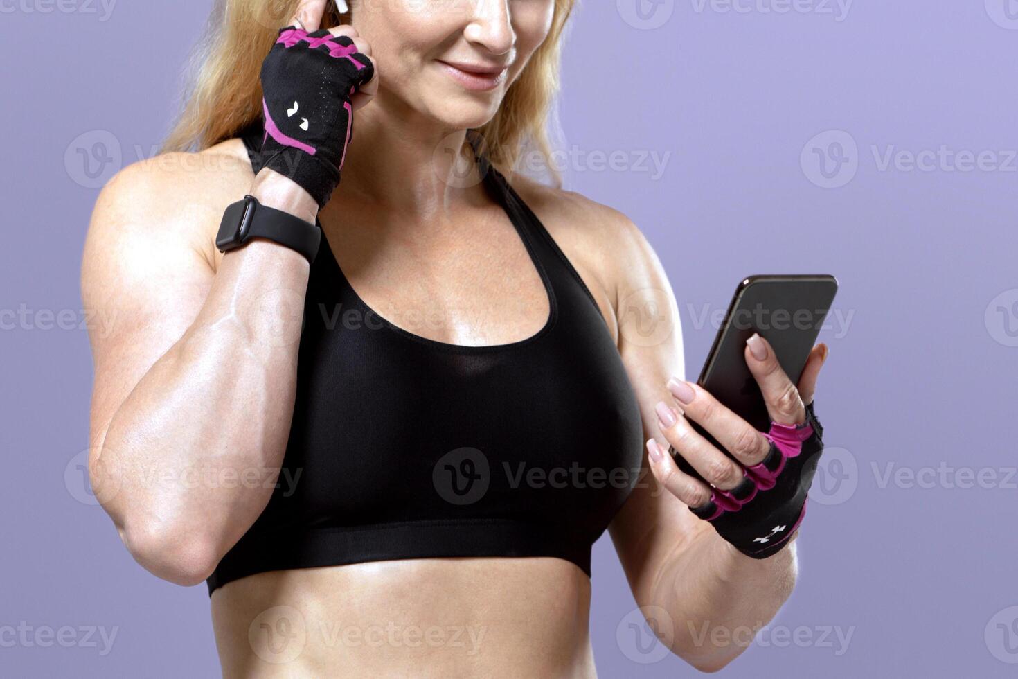 Favorite music for workout at home or gym at lockdown. Happy muscular middle aged lady coach in sportswear with gloves looks in smartphone and puts on wireless earphone, isolated on violet background photo
