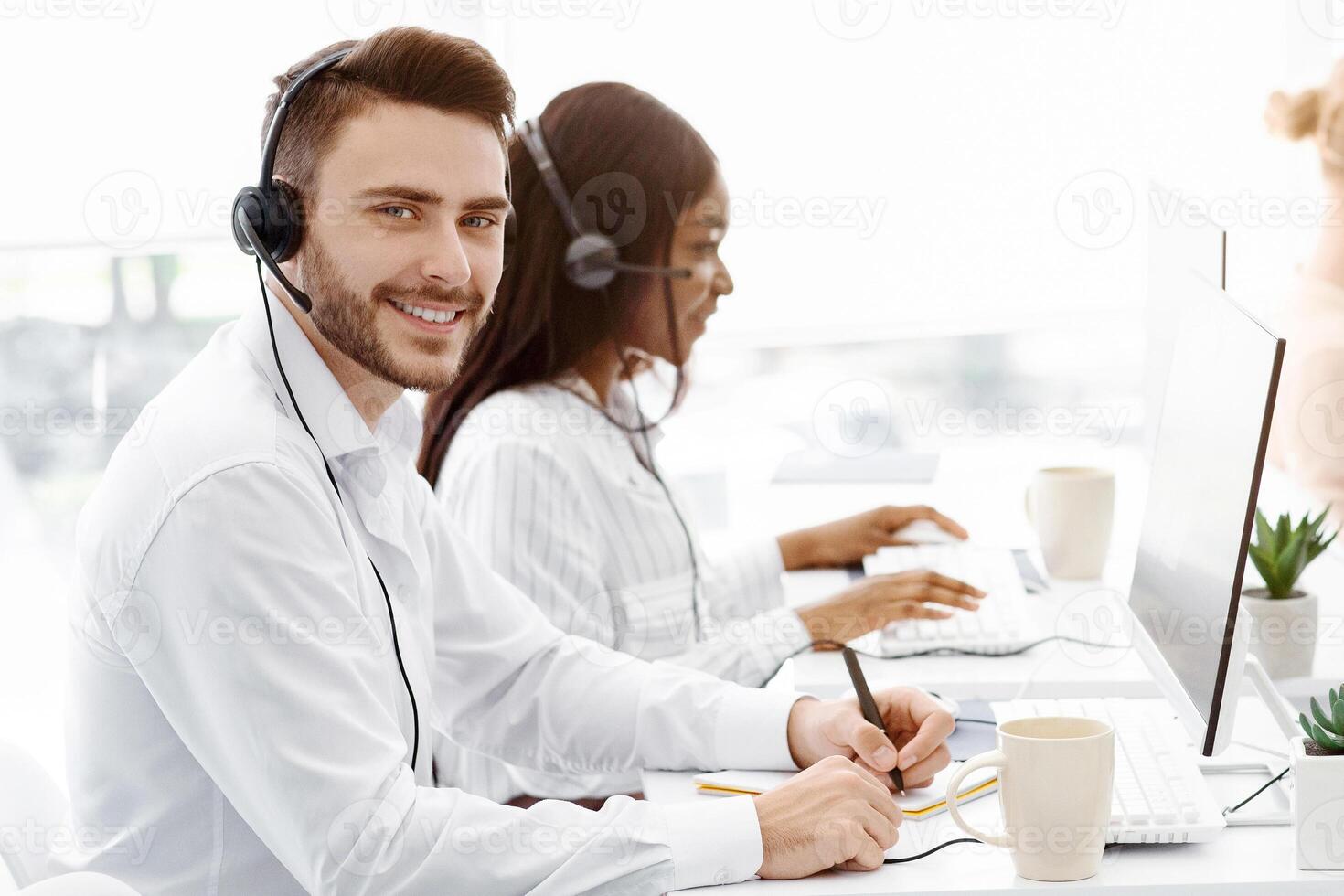 Handsome call center operator smiling at camera and his coworkers talking to clients at office, copy space photo