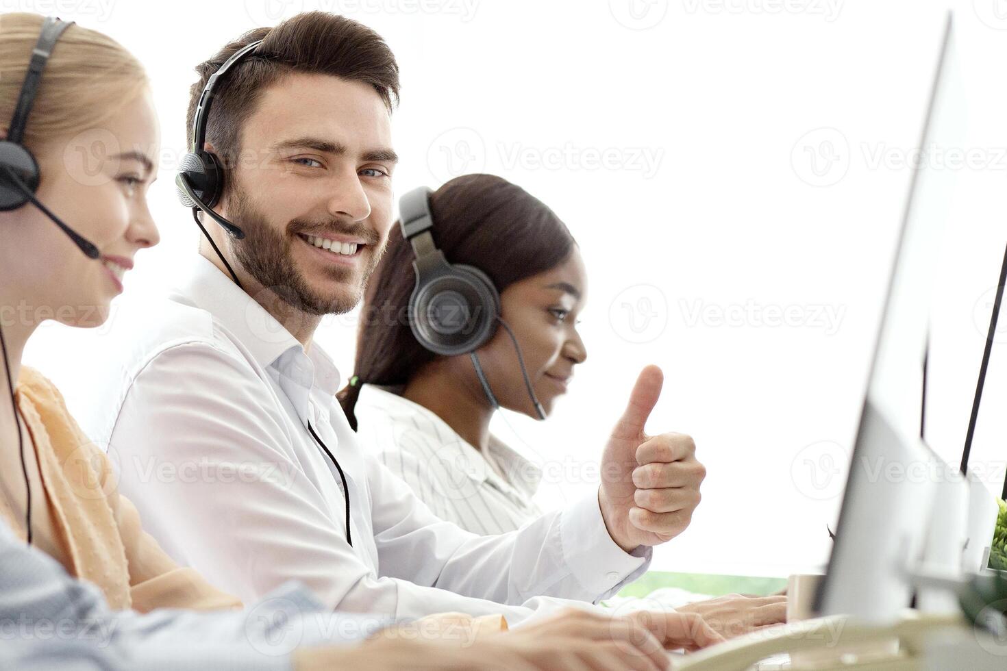 Happy male call centre operator showing thumb up gesture, working with his colleages at modern office. Positive tech support consultant providing telemarketing services photo