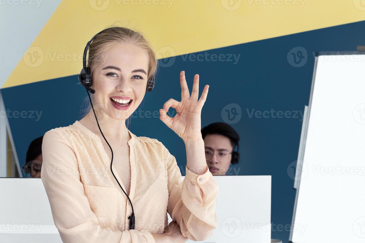 Beautiful female helpline consultant looking at camera and showing OKAY gesture in modern office. Call centre operator expressing positivity and her collagues at open workspace photo