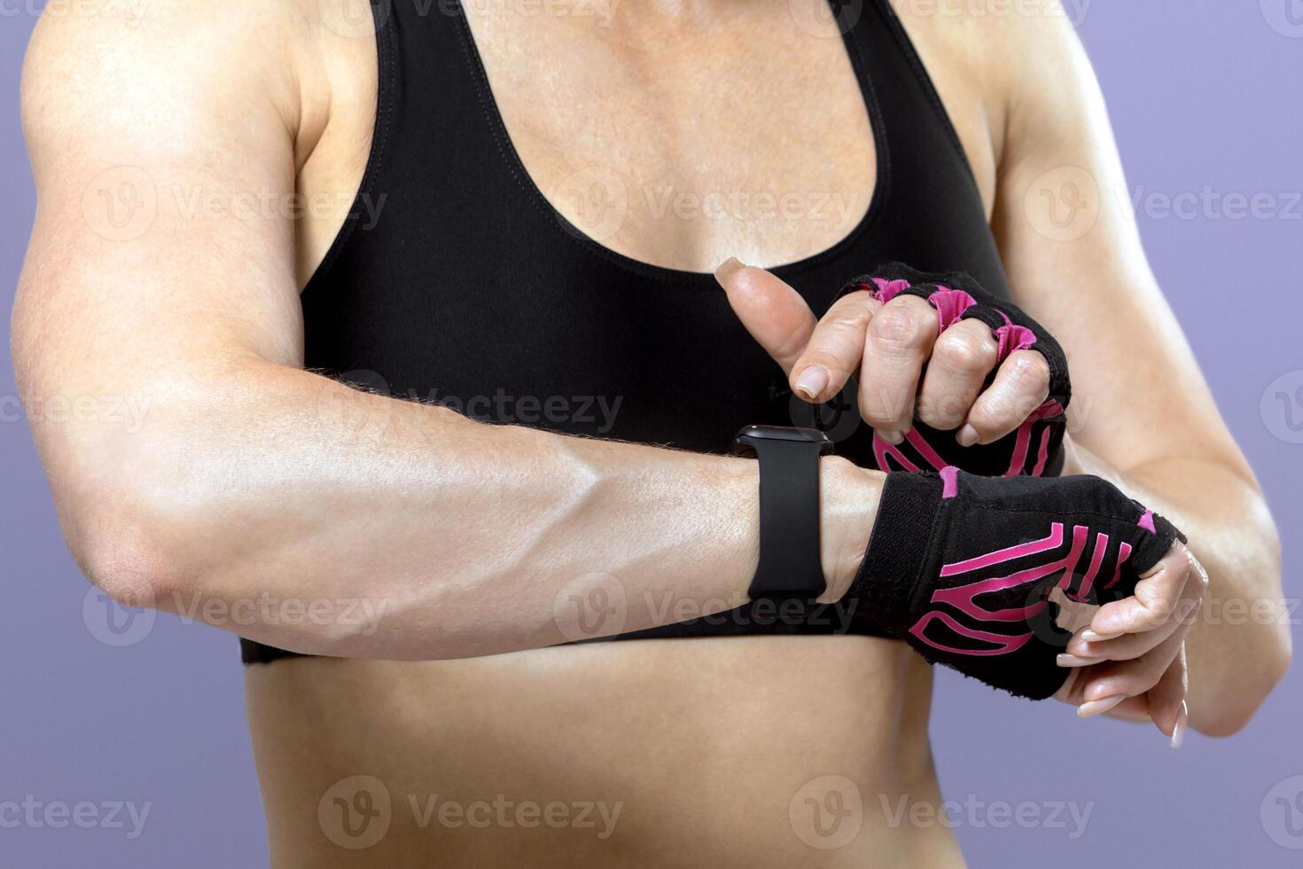 Modern gadgets and bodybuilder coach checks heartbeat. Serious adult tired muscular female in sportswear with gloves and fitness tracker, isolated on purple background, free space, studio shot photo