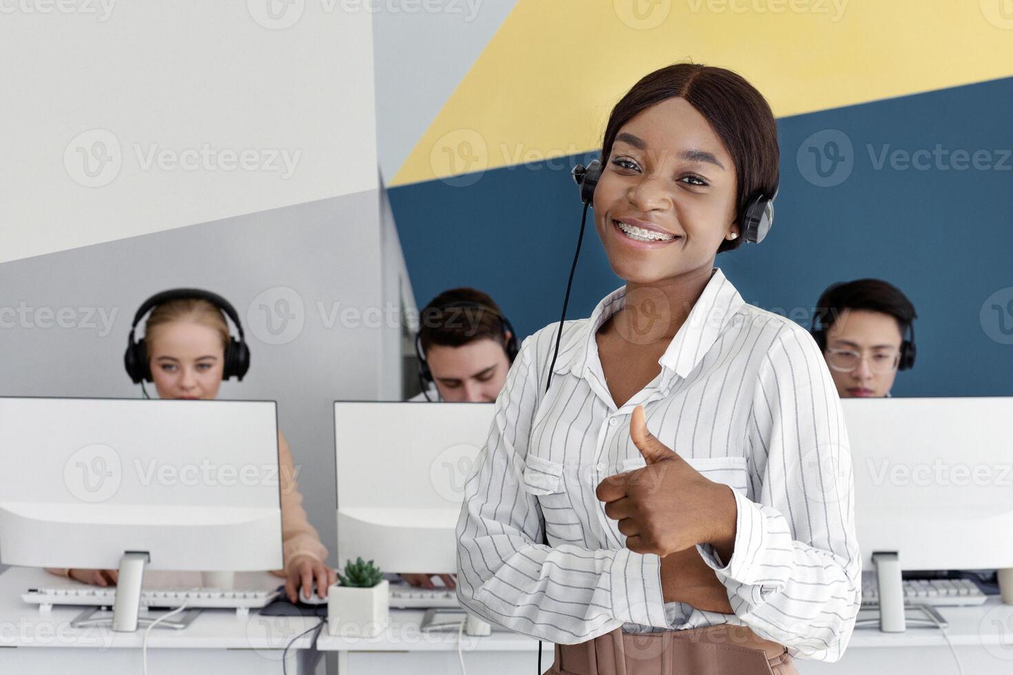 Black female hotline operator smiling and showing thumb up at modern call centre. Customer support agent expressing approval, and her colleagues working at open space office photo
