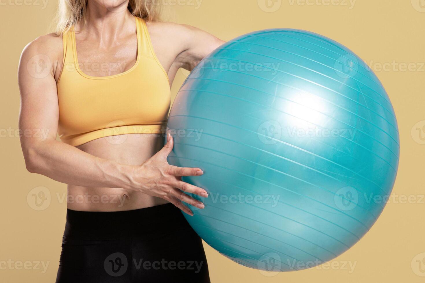 Exercise with fitness ball and muscular body. Friendly muscular adult female in sports uniform holding workout equipment, isolated on beige background, empty space, cropped, panorama, studio shot photo