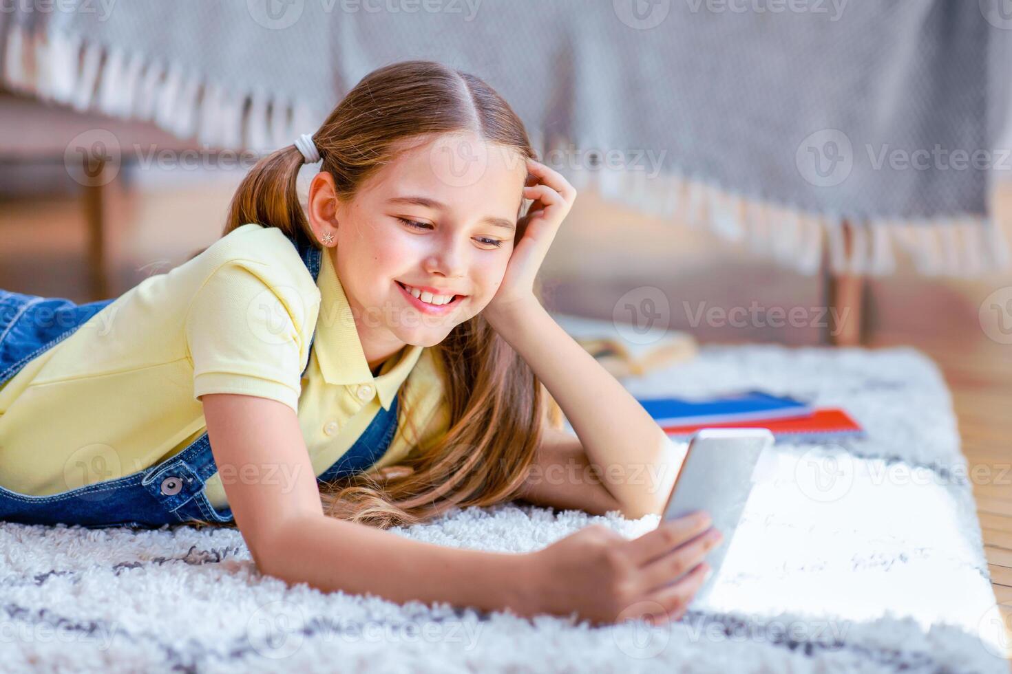 Amazing Application. Portrait of positive glad teenage kid with ponytails using smart phone, chatting, typing and sending message, watching funny , lying on the floor carpet at home photo