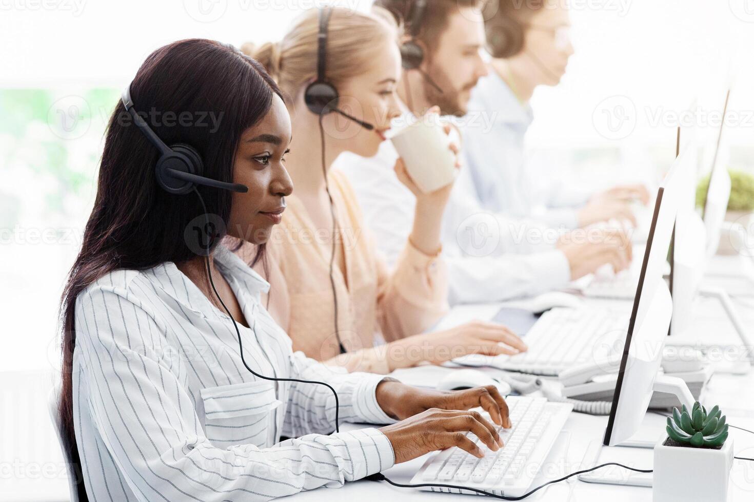 Line of call centre employees with headphones working on computers at open space office photo