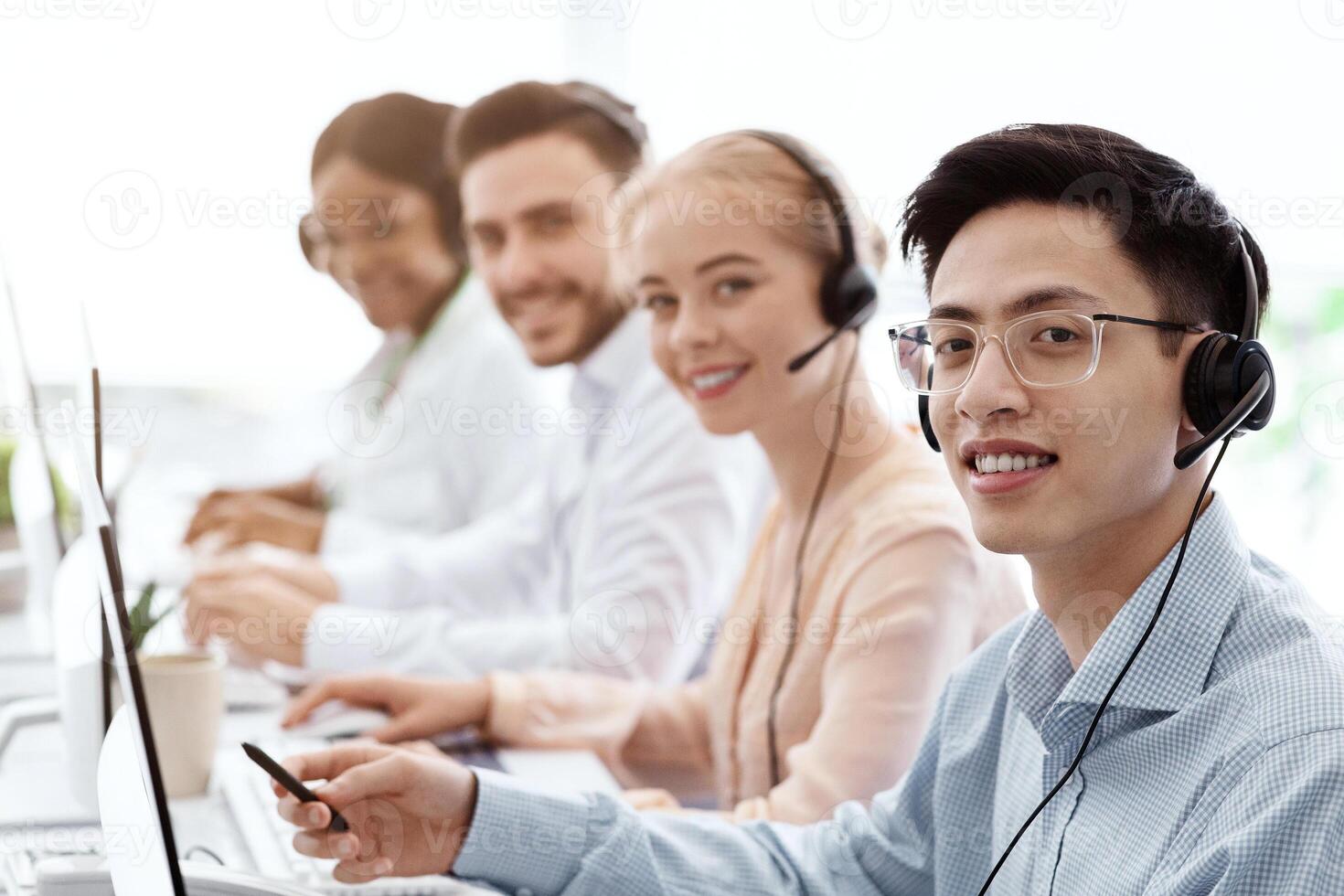 Smiling call center workers sitting in row near their computers at light office, empty space photo