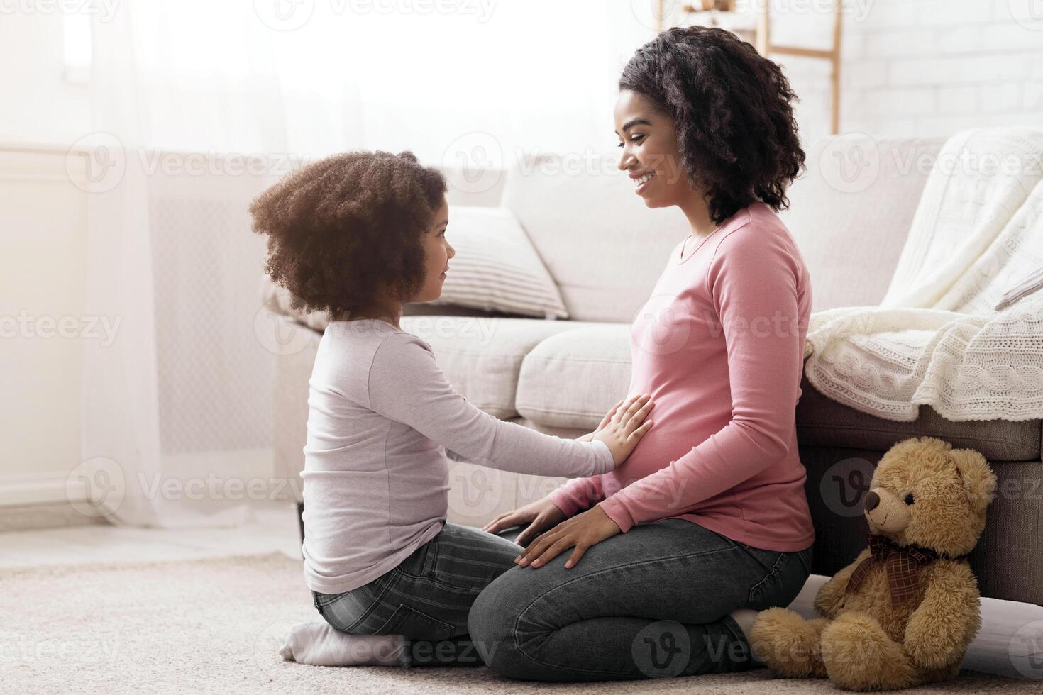 Family Connection. Adorable little daughter touching pregnant mom's belly while playing together on floor in living room, side view with free space photo