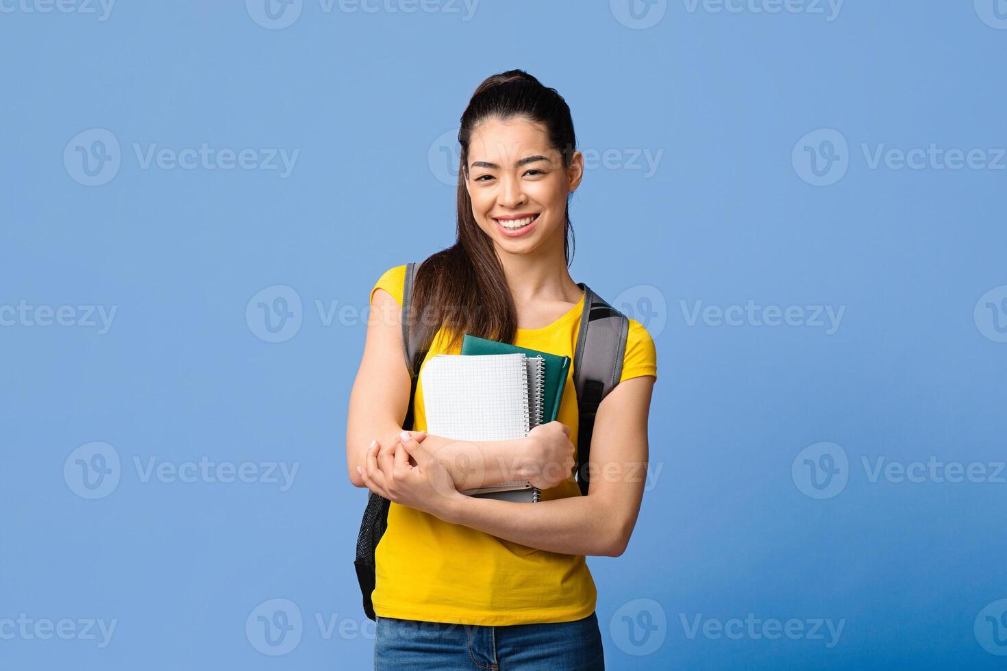 Portraif of smiling asian student girl with backpack and workbooks posing over purple studio background, looking at camera, copy space photo