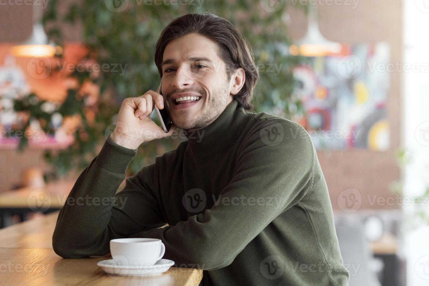 Good looking young man cheerfully talking on smartphone at lounge cafe, free space photo