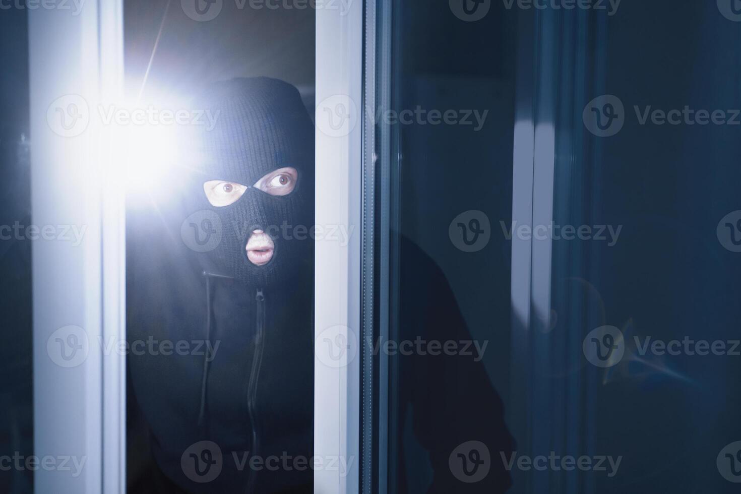 Robbery Concept. Aware masked villain lurking into house through window or glass door, using torch in the dark photo