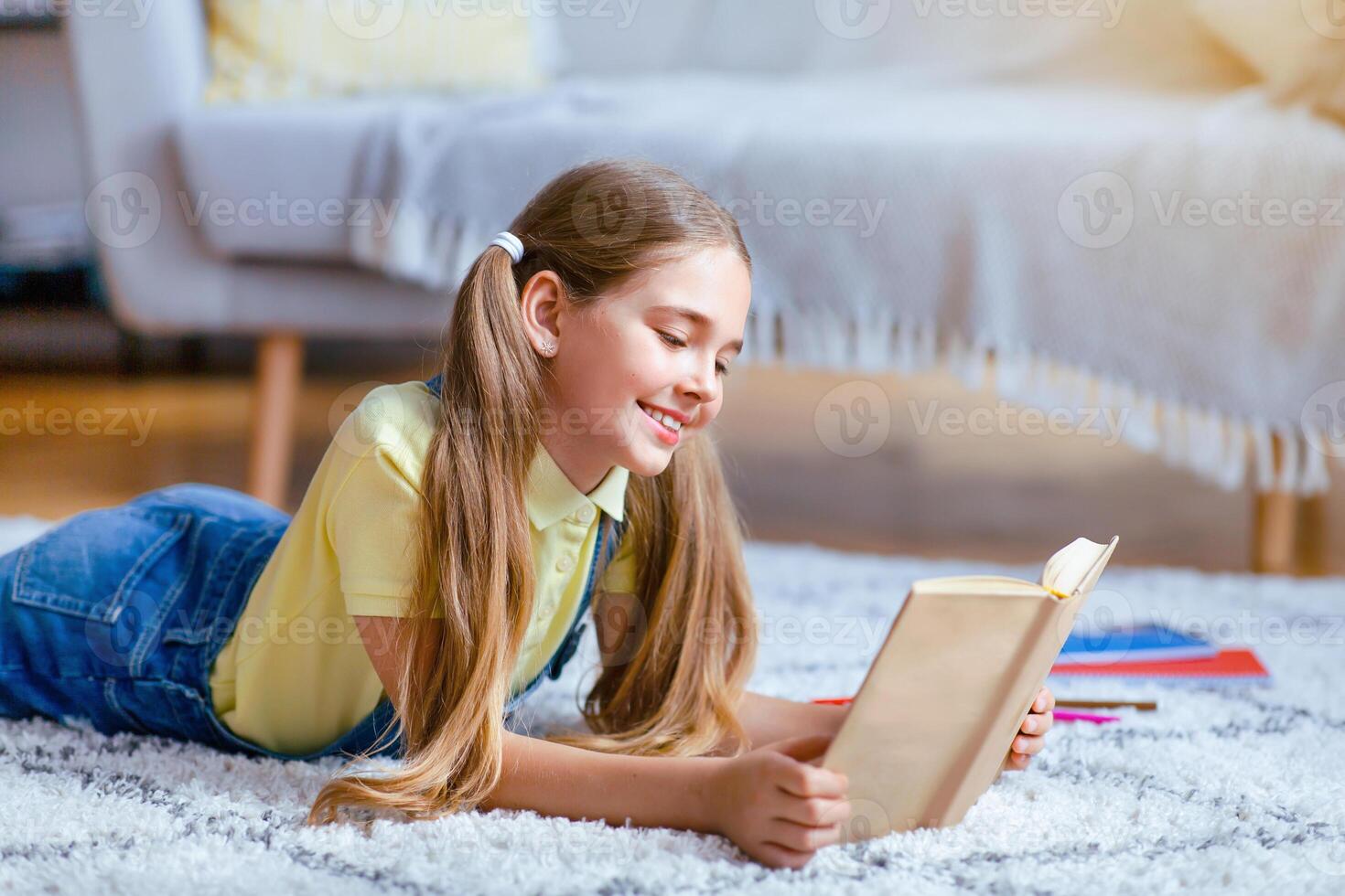 Leisure Concept. Portrait of cute smiling teenage girl lying on the floor carpet at home in living room, holding and reading paper book, enjoying story, copy space. Pupil resting and relaxing photo