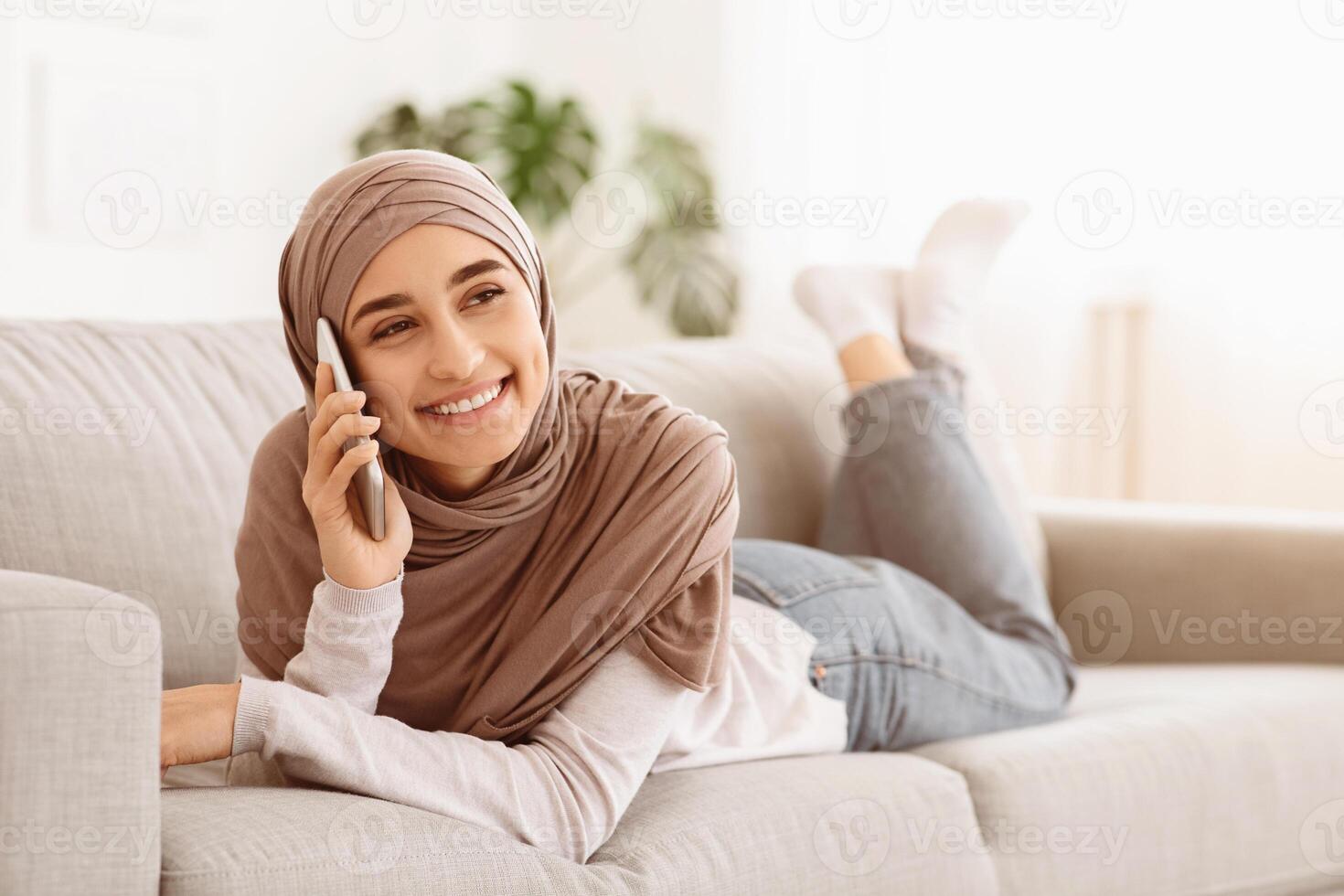Smiling Arabic Girl Relaxing On Couch At Home, Talking On Cellphone, Chatting With Friends, Lying On Comfortable Sofa In Living Room, Free Space photo