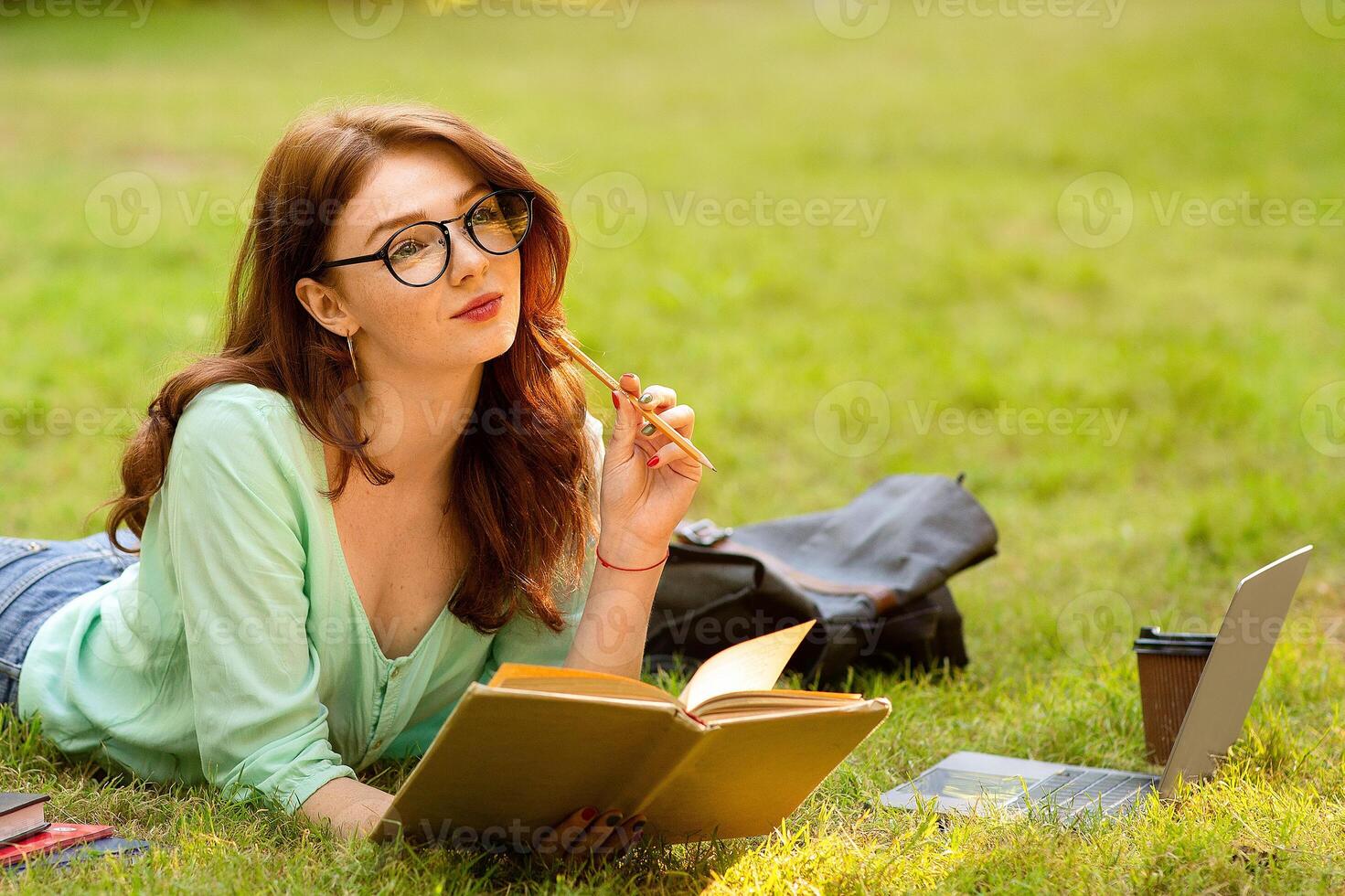 Portrait Of Dreamy Student Girl In Eyeglasses Studying Outdoors, Reading Book While Relaxing On Lawn In Park, Free Space photo