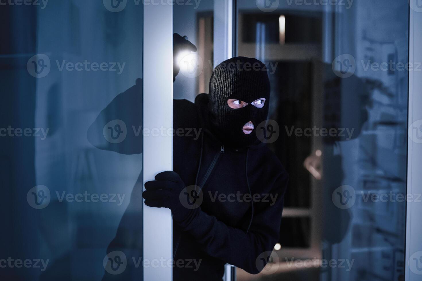 Intrusion Concept. Attentive looter with black hat sneaking into house through open glass door, searching with flashlight photo