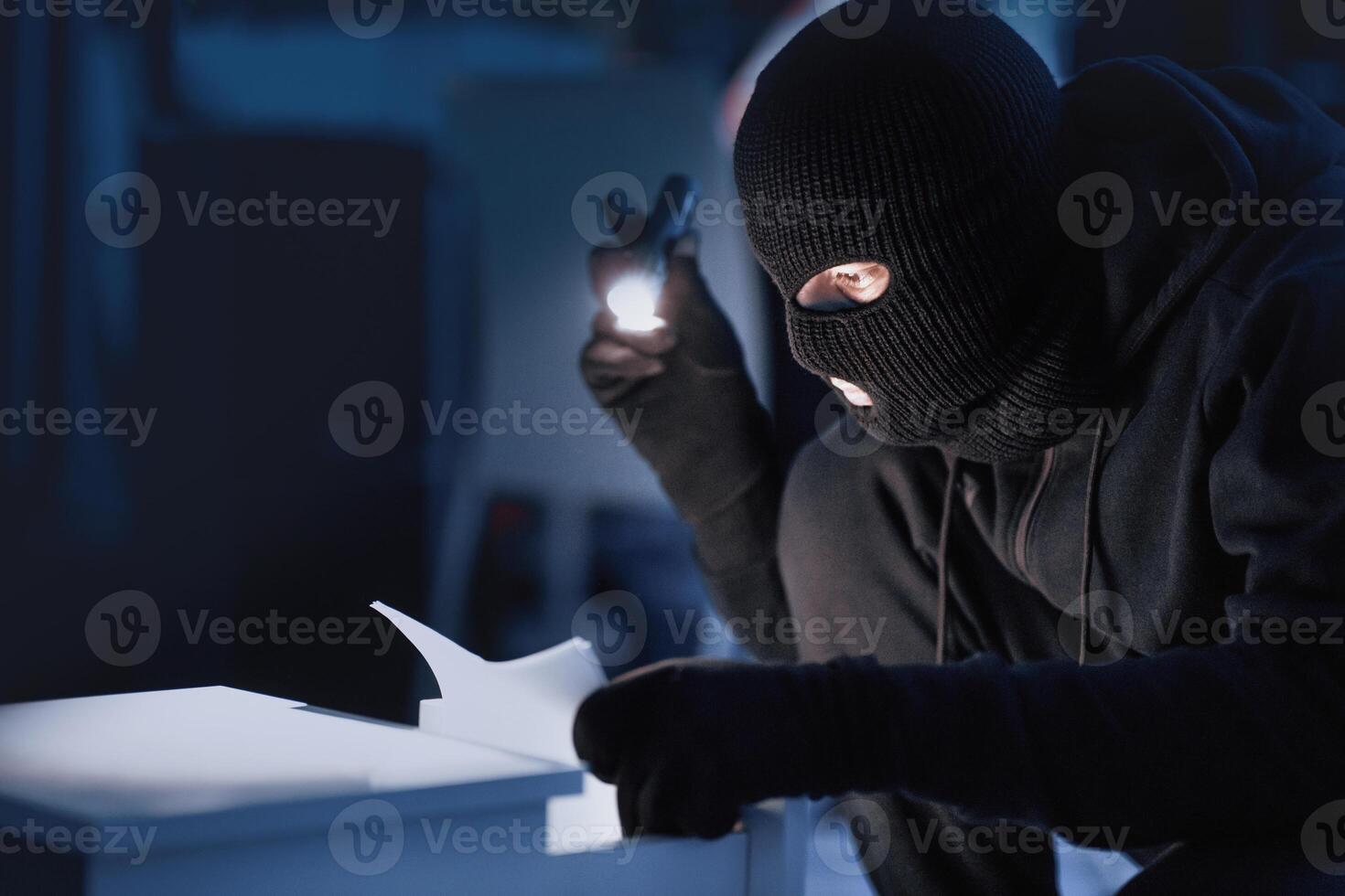 Misappropriation Concept. Masked spy holding flash light and checking papers in office drawer in the dark photo