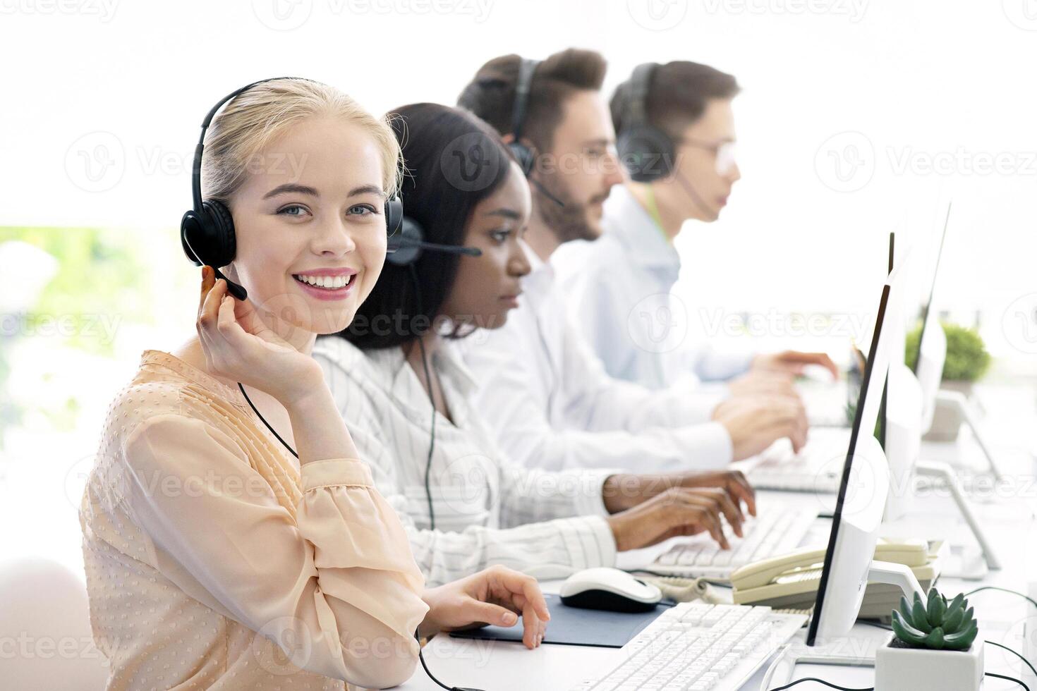 Team of diverse hotline operators with headphones providing service to clients at call center. Young customer support representatives working in telemarketing or telecommunications at modern office photo