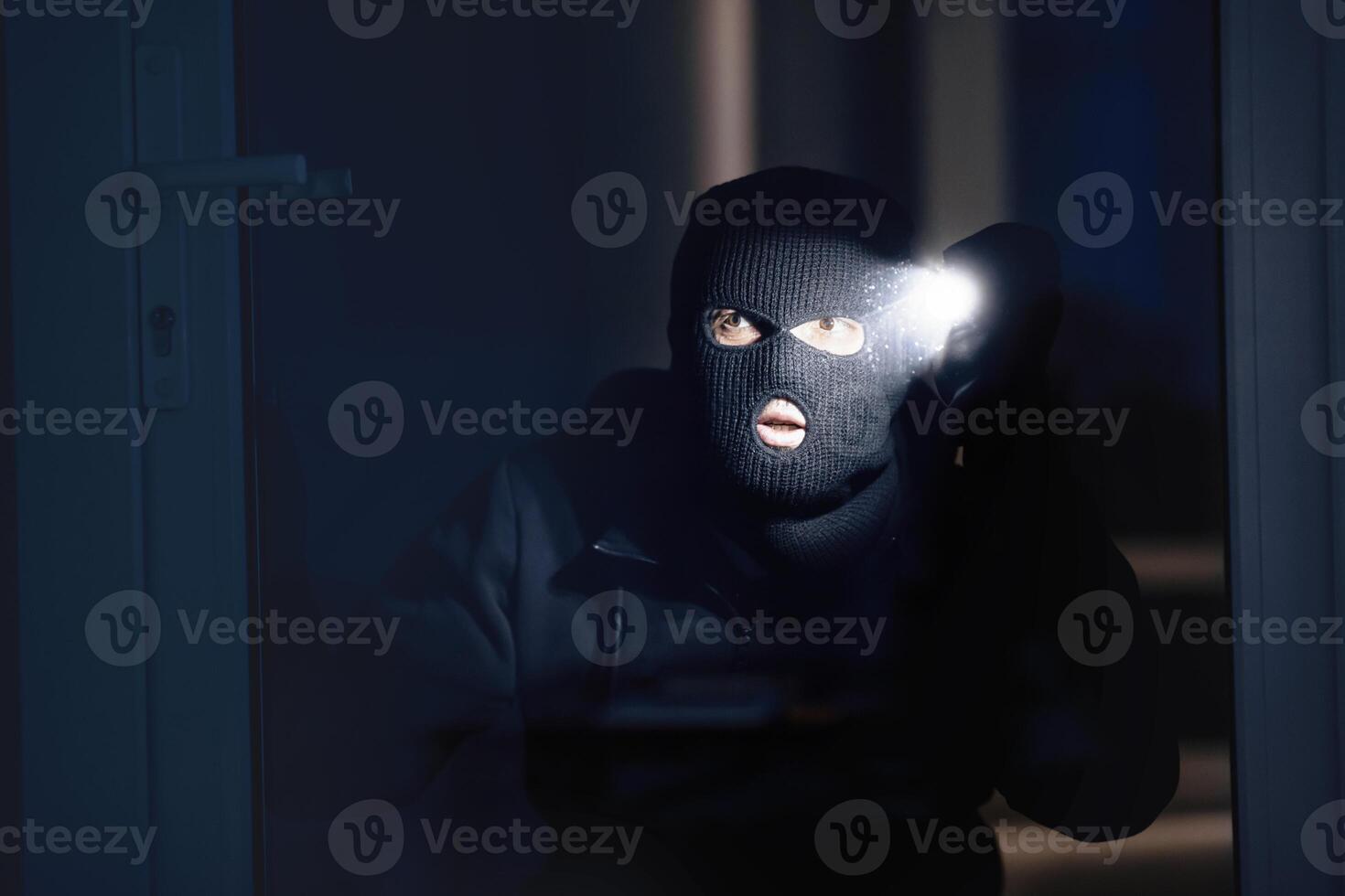 Shadowing Concept. Criminal wearing black balaclava watching you through window or door, using flashlight torch photo