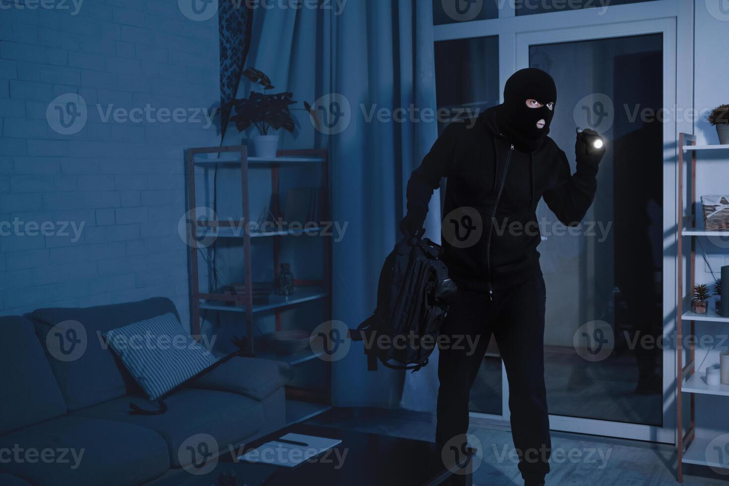 Robbery Concept. Disguised burglar standing in apartment or office, using flashlight, searching something to steal photo