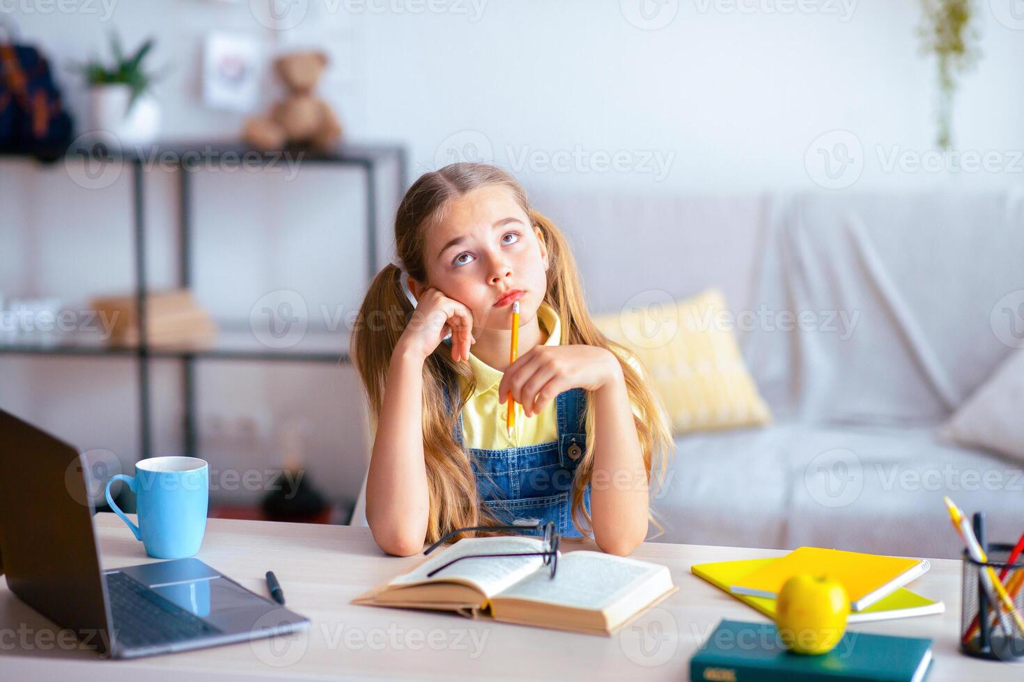 Portrait of pensive pupil sitting at desk with open book and doing homework, looking up, copy space photo