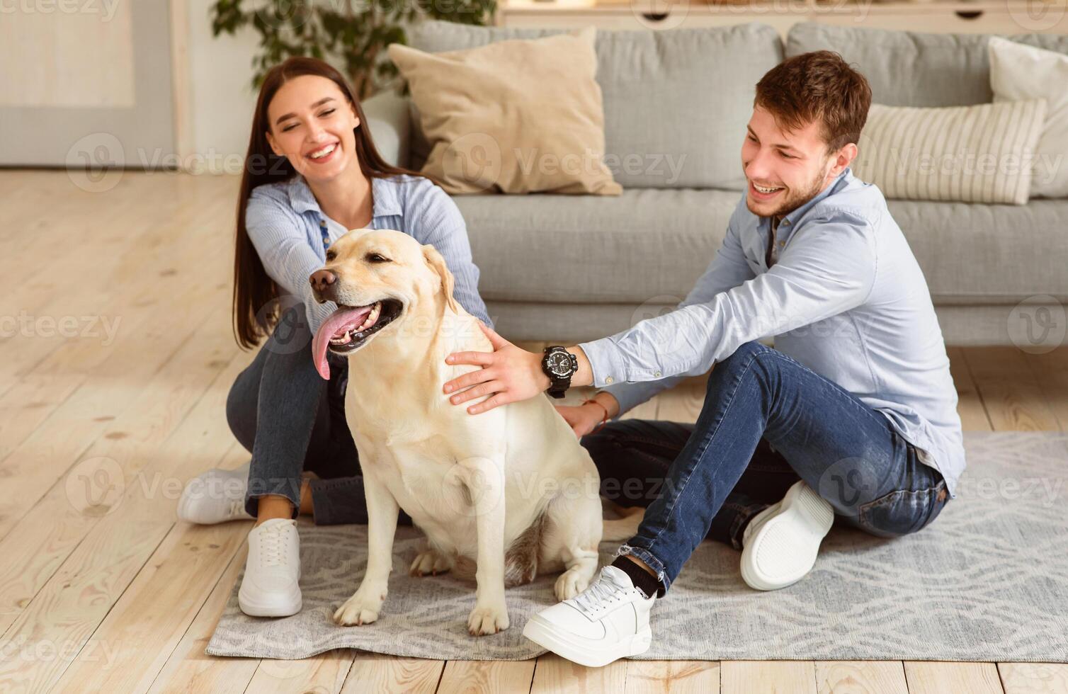 Happy Loving Family Concept. Portrait of cheerful spouses sitting on the floor in modern apartment, spending time together with dog. Man and woman owners playing with labrador pet at home photo