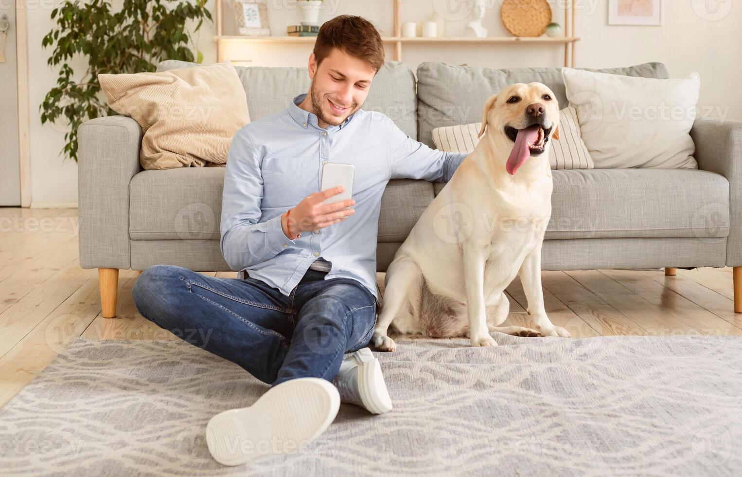 Lifestyle Concept. Portrait of smiling man using smartphone sitting on the carpet with his labrador, stroking dog. Guy spending time at home, chatting at social media, texting sms or reading message photo