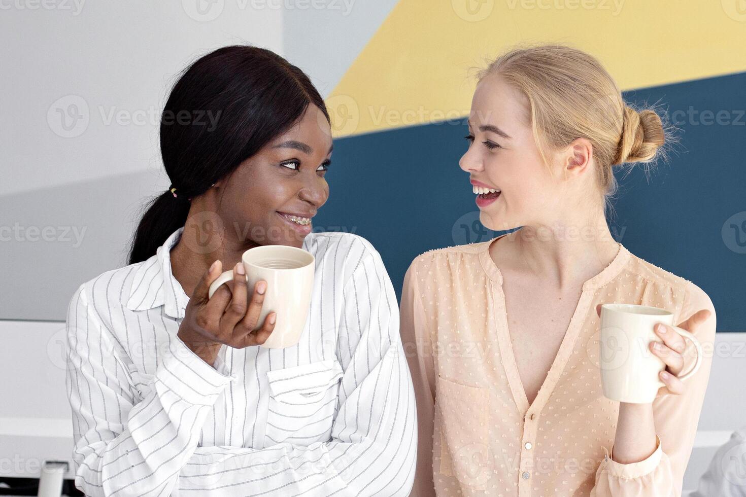 Diverse customer support operators on coffee break at modern call centre, space for text. Female telemarketing specialists with hot drinks making small talk at open space office photo