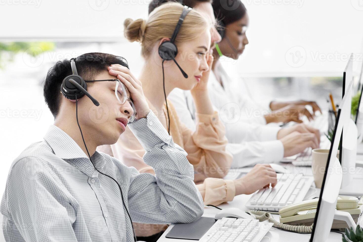 Overworked male hotline agent sleeping in front of computer monitor at call centre office, copy space. Exhausted Asian customer support manager napping at workplace near his colleagues photo