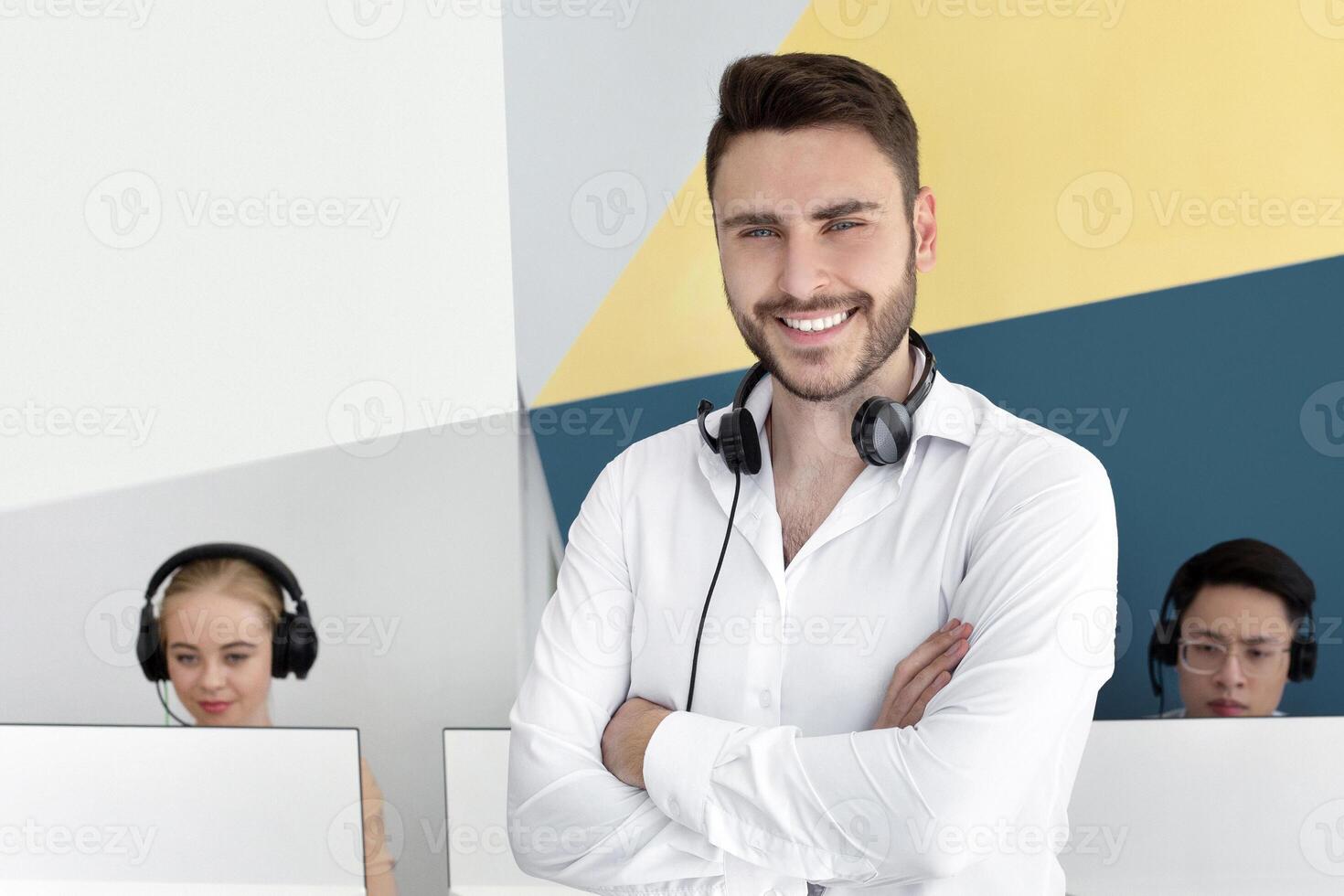 Portrait of positive male customer support representative and his colleagues at contemporary call centre. Happy hotline operator with headset looking and camera and smiling at office photo