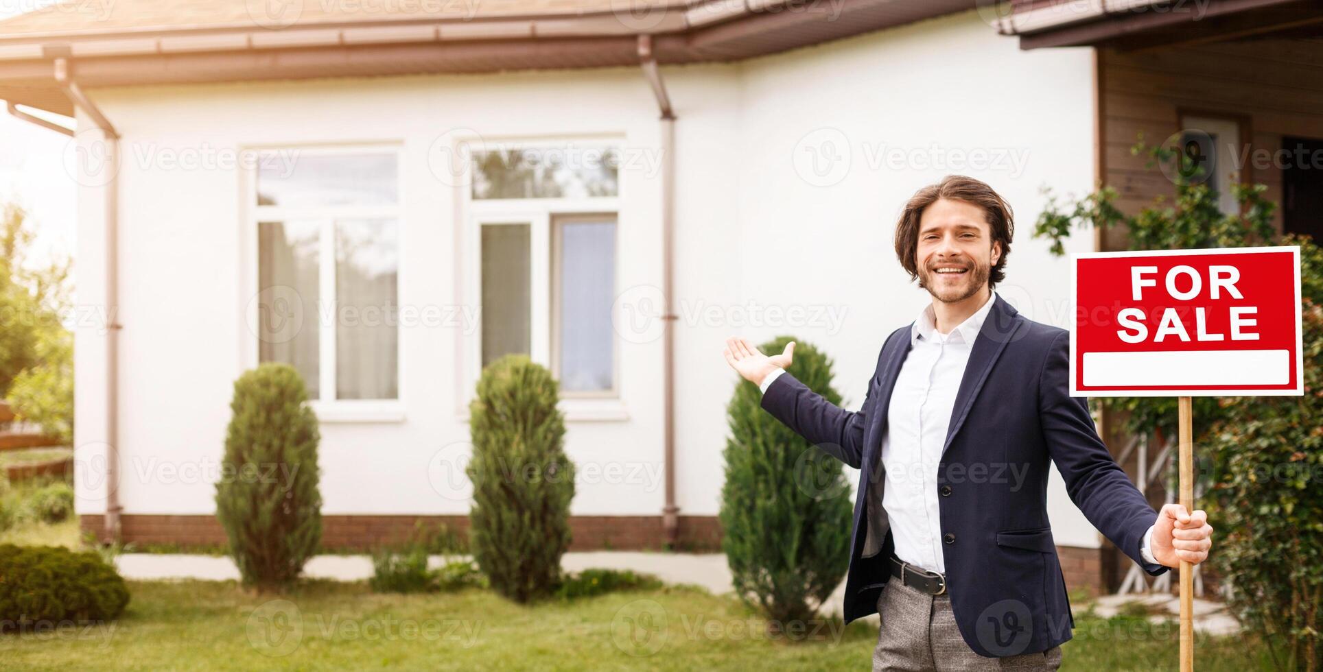 Cheerful real estate agent with FOR SALE sign pointing towards beautiful house, outdoors. Copy space photo