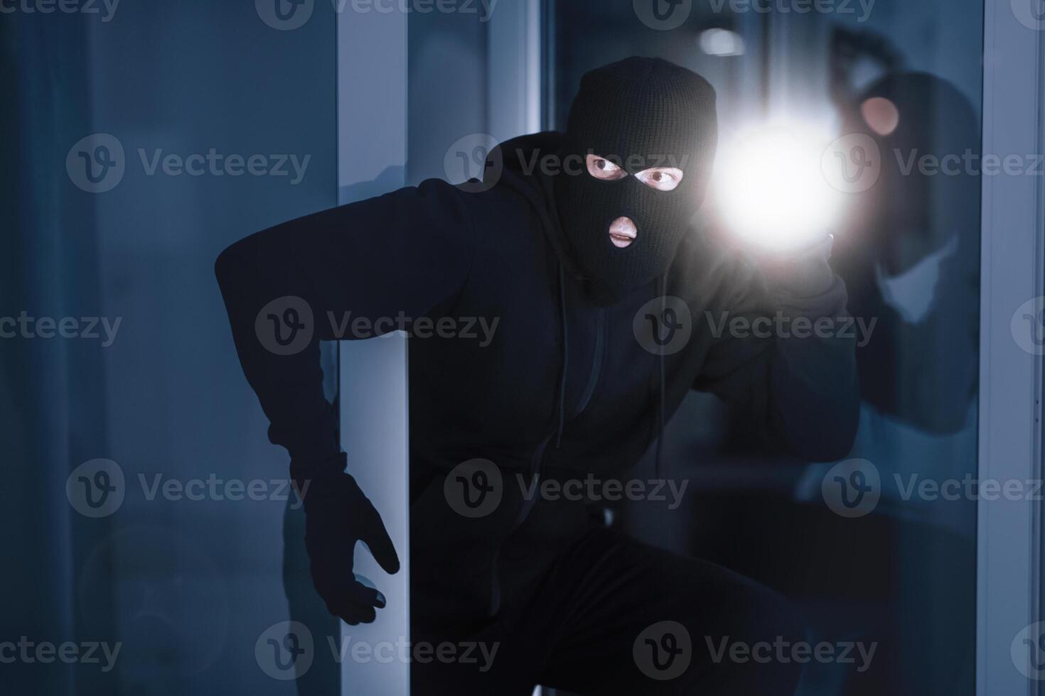Time For Crime. Cautious burglar looking and peeping into building through window or glass door with torch photo