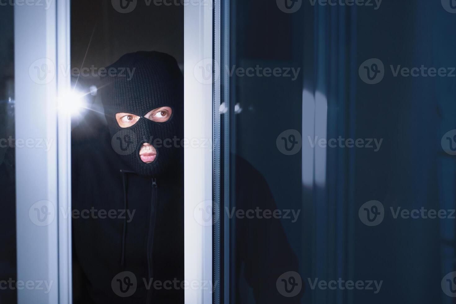 Robbery Concept. Disguised burglar looking through glass in an apartment or office to steal something, using flashlight photo