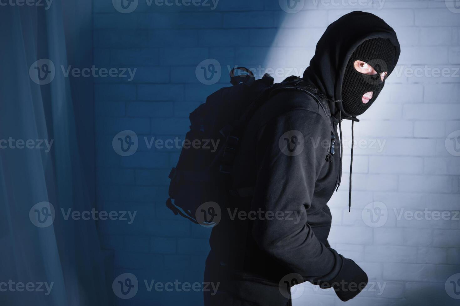Masked robber sneaking into apartment or office at night, wearing hoodie sweatshirt and backpack, copy space photo