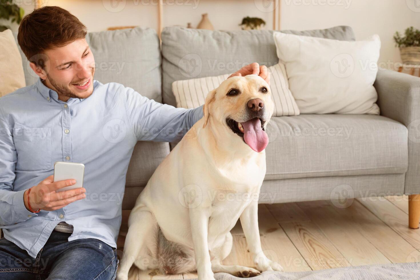 Animal Love And Care Concept. Happy guy holding mobile phone sitting on the floor with dog and fondling his pet. Portrait of male owner spending time at home in living room, scratching doggy photo